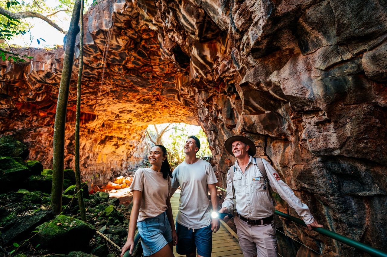 Three people are walking through a rocky cave with an opening. They are smiling and dressed in casual clothing, with one person holding a flashlight.