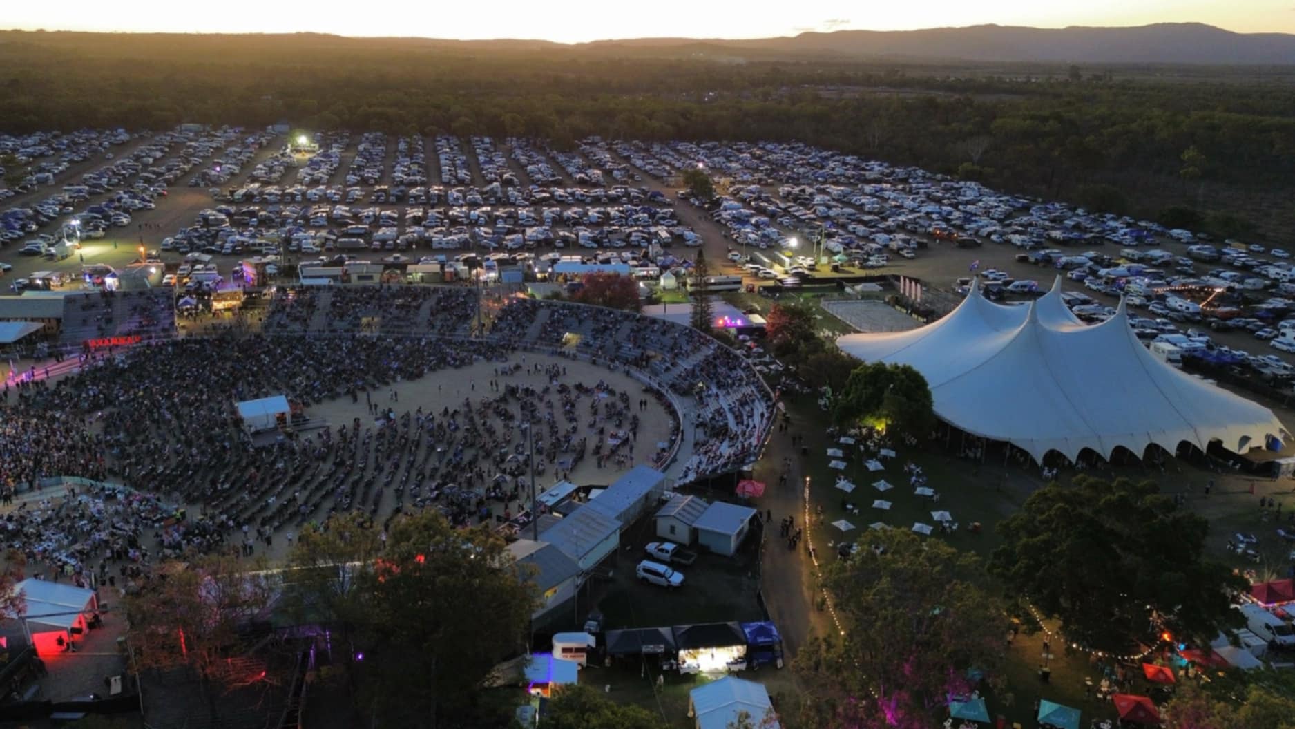 Aerial view of a large outdoor concert with a circular seating area and a white tent. Cars fill the parking lot in the background. The scene is set at sunset.