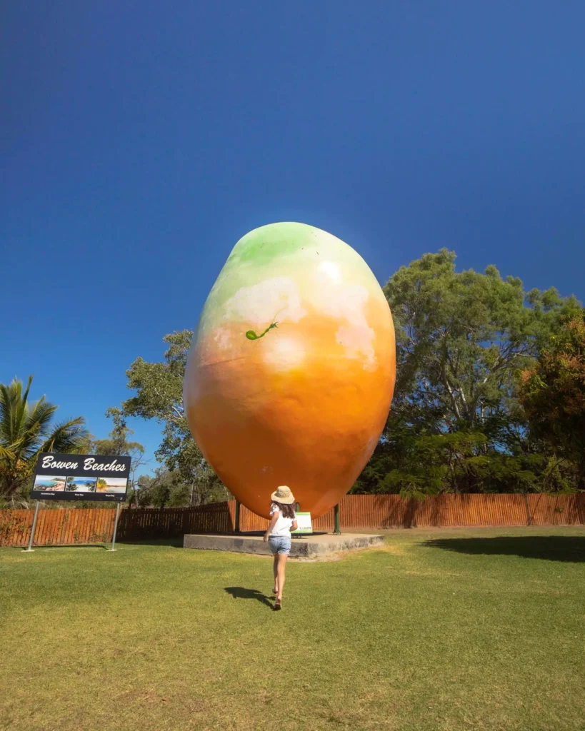 A person stands facing a large, mango-shaped sculpture in a grassy area with trees and a clear blue sky in the background. A sign that reads 