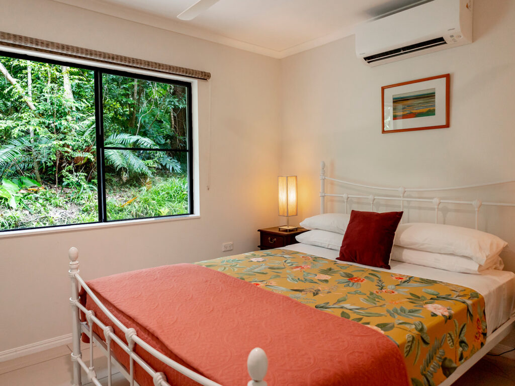 A cozy bedroom with a white metal bed, floral bedding, and a red blanket. A window shows a garden view. A central lamp and a wall-mounted air conditioner are visible.