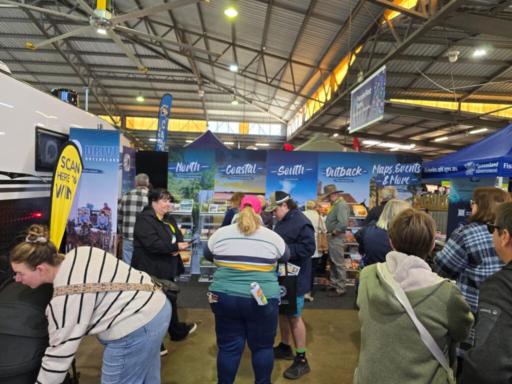 People visit various informational booths at an indoor exhibition, engaging with staff and looking at brochures and displays under blue banners promoting different regions.