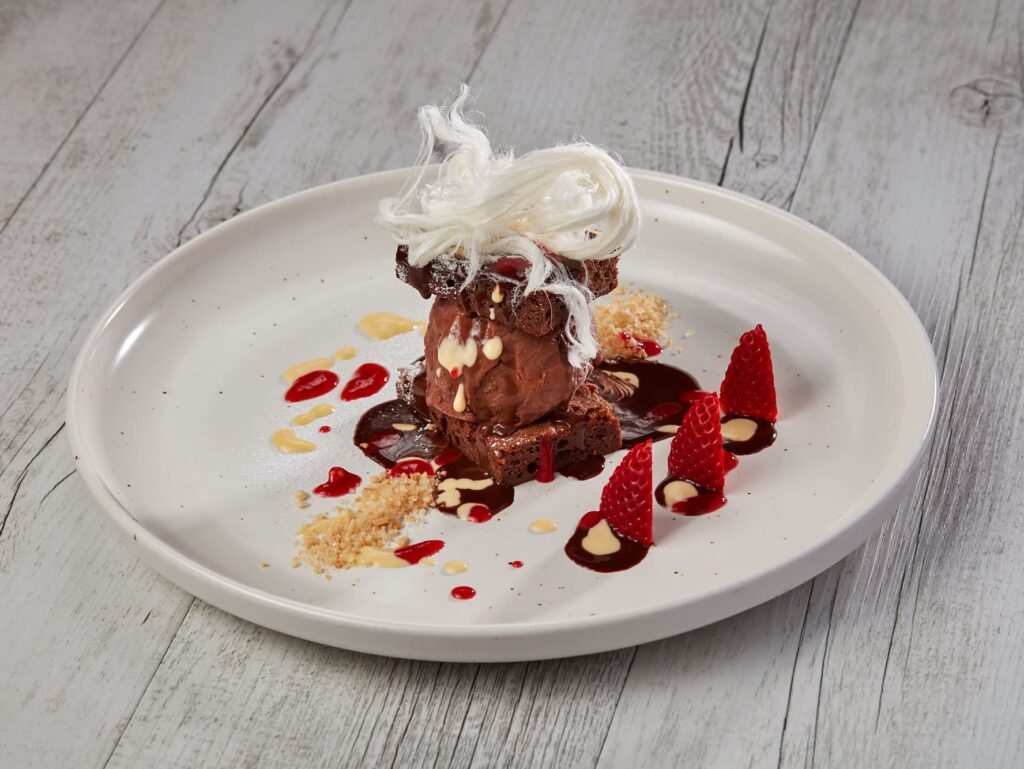 A plated chocolate dessert with spun sugar on top, surrounded by strawberry halves, berry sauce, cream drizzle, and a sprinkle of crumbs on a white plate.
