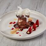 A plated chocolate dessert with spun sugar on top, surrounded by strawberry halves, berry sauce, cream drizzle, and a sprinkle of crumbs on a white plate.