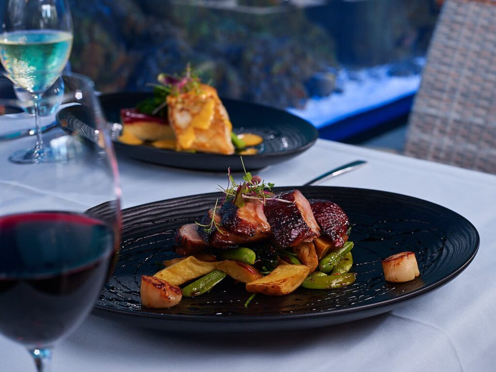 A plated gourmet meal with roasted meat, potatoes, and vegetables on a black plate, accompanied by glasses of red and white wine on a white tablecloth.