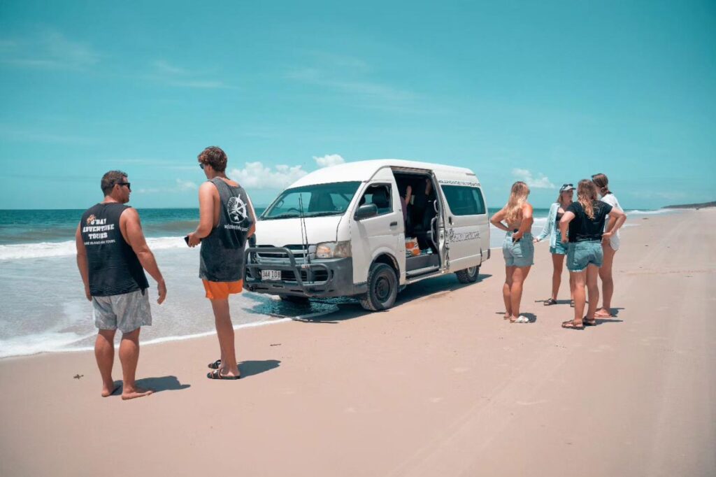 A group of people stands near a white van stuck in the sand on a sunny beach, with the ocean and blue sky in the background.