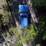 A blue car drives along a narrow dirt road surrounded by green trees, seen from directly above.