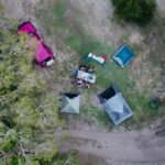 Aerial view of a campsite with people seated at a table, two tents, a pink vehicle, and surrounding trees on grassy and dirt terrain.