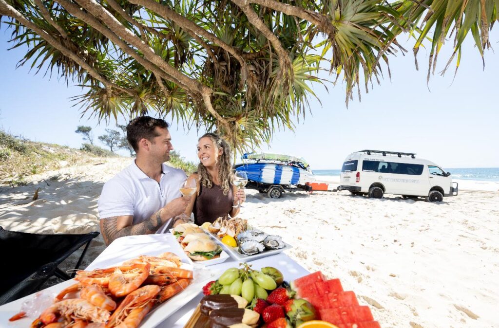 A man and woman sit at a table on a beach under a tree, enjoying seafood and fruit; a van and surfboards are parked nearby, with the ocean in the background.