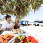 A man and woman sit at a table on a beach under a tree, enjoying seafood and fruit; a van and surfboards are parked nearby, with the ocean in the background.
