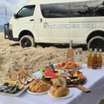 A table set with seafood, sandwiches, fruit, and drinks on a sandy beach, with a white tour van parked in the background under a partly cloudy sky.