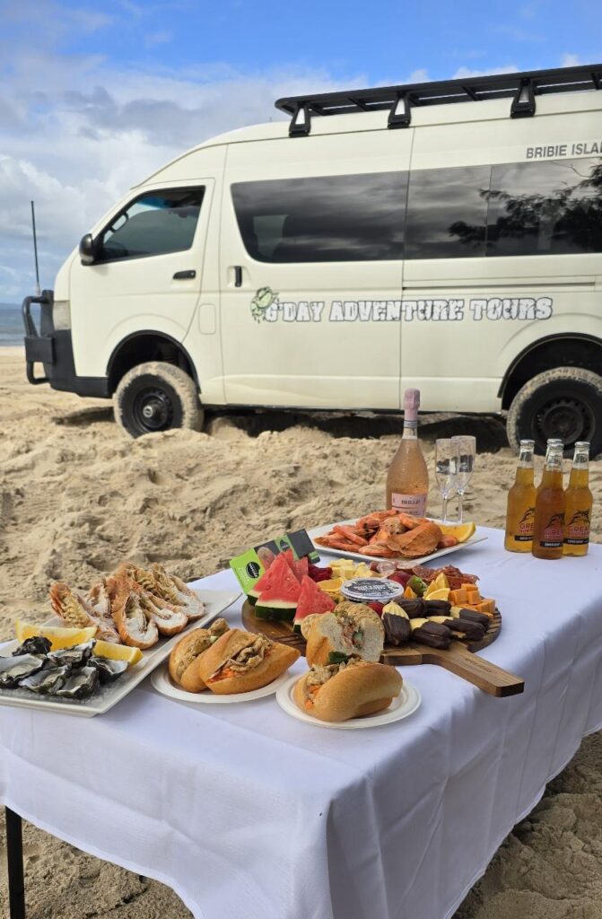 A table set with seafood, sandwiches, fruit, and drinks on a sandy beach, with a white tour van parked in the background under a partly cloudy sky.