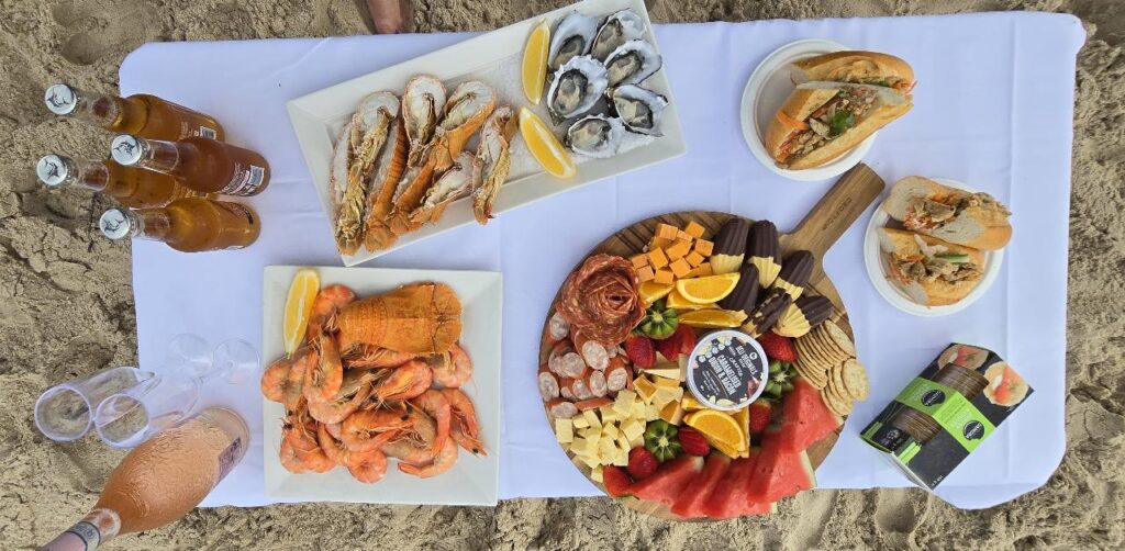 A picnic setup on sand with seafood, oysters, shrimp, assorted cheeses, charcuterie, bread rolls, a bottle of rosé, beers, and glasses on a white cloth.
