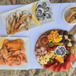 A picnic setup on sand with seafood, oysters, shrimp, assorted cheeses, charcuterie, bread rolls, a bottle of rosé, beers, and glasses on a white cloth.