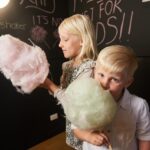 Two young children stand indoors holding large sticks of cotton candy, with "fairy floss" and other words written in pink chalk on a black wall behind them.