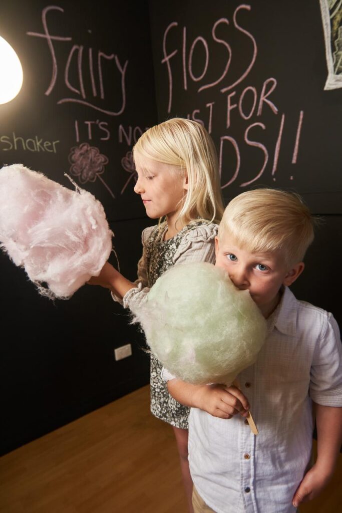 Two young children stand indoors holding large sticks of cotton candy, with "fairy floss" and other words written in pink chalk on a black wall behind them.