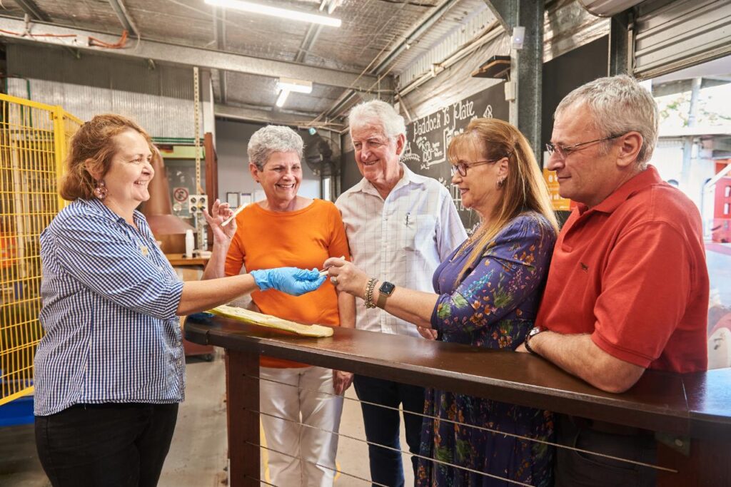 Five adults stand indoors near a railing; one woman wearing gloves hands an item to another woman while the group looks on and smiles.