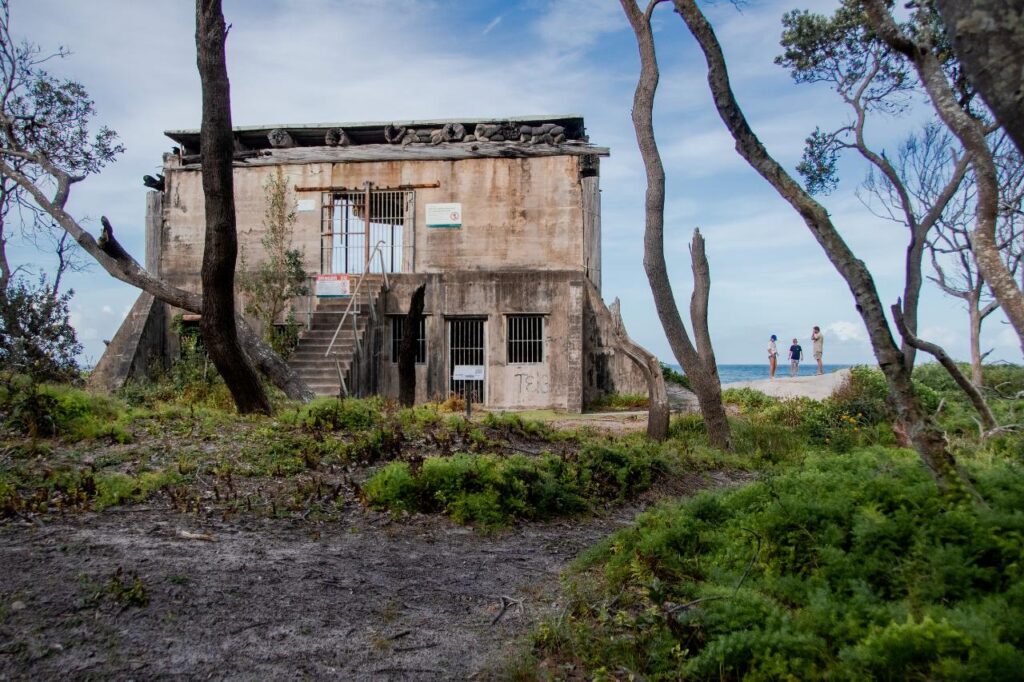 An old, weathered concrete building with barred windows stands among trees near the coast; three people walk on a path in the background.