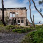 An old, weathered concrete building with barred windows stands among trees near the coast; three people walk on a path in the background.