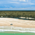 A white SUV is parked on a sandy beach near the shoreline, with a narrow river and dense forest in the background under a partly cloudy sky.