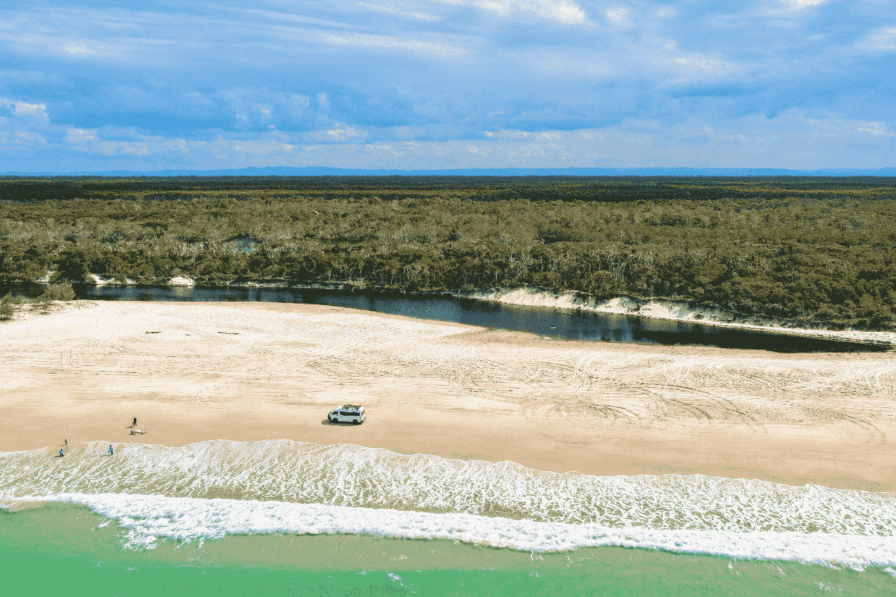A white SUV is parked on a sandy beach near the shoreline, with a narrow river and dense forest in the background under a partly cloudy sky.