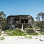 Abandoned concrete bunker with weathered walls sits partially hidden by trees at the edge of a sandy beach under a clear blue sky.