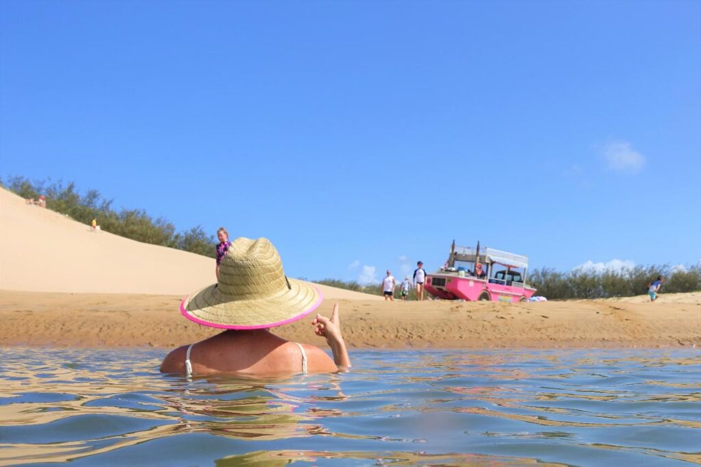 Person in a straw hat wades in shallow water near a sandy shore, facing a pink boat and several people under a clear blue sky.