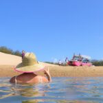 Person in a straw hat wades in shallow water near a sandy shore, facing a pink boat and several people under a clear blue sky.