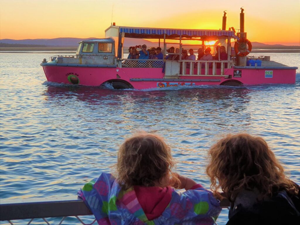 Two children watch a pink boat with passengers on the water at sunset, with mountains visible in the background.
