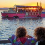 Two children watch a pink boat with passengers on the water at sunset, with mountains visible in the background.
