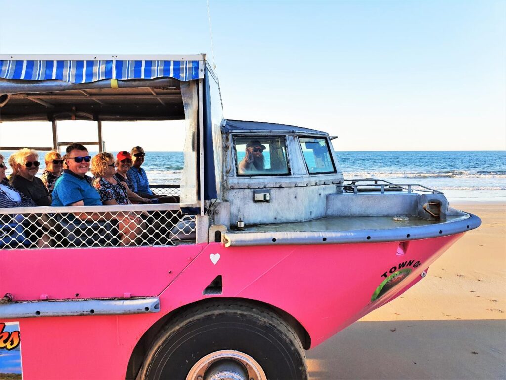 A group of people sit in an amphibious tour vehicle driving on a sandy beach next to the ocean under a clear sky.