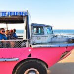 A group of people sit in an amphibious tour vehicle driving on a sandy beach next to the ocean under a clear sky.