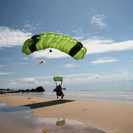 A person paragliding lands on a sandy beach near the shoreline under a partly cloudy sky.