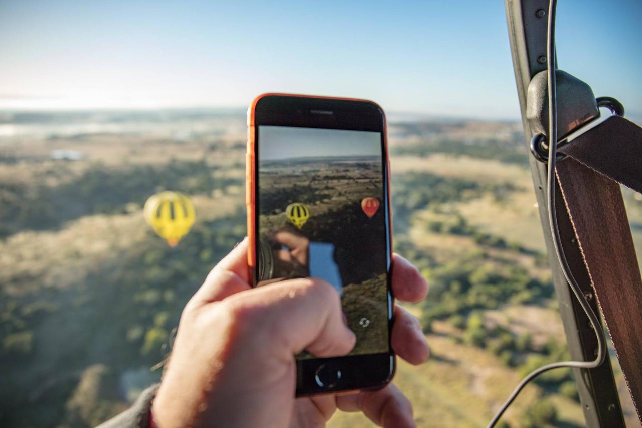 A person holds a smartphone, taking a photo of hot air balloons flying over a landscape from inside another hot air balloon.