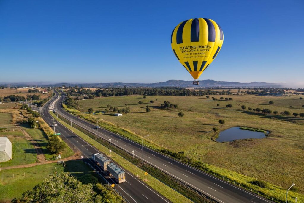 A yellow and black hot air balloon floats above a rural landscape with open fields, a pond, and a highway with vehicles under a clear blue sky.