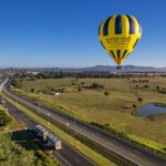 A yellow and black hot air balloon floats above a rural landscape with open fields, a pond, and a highway with vehicles under a clear blue sky.