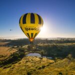 A yellow and black hot air balloon with "discover Ipswich" printed on it floats above a rural landscape at sunrise; a small helicopter is visible in the distance.