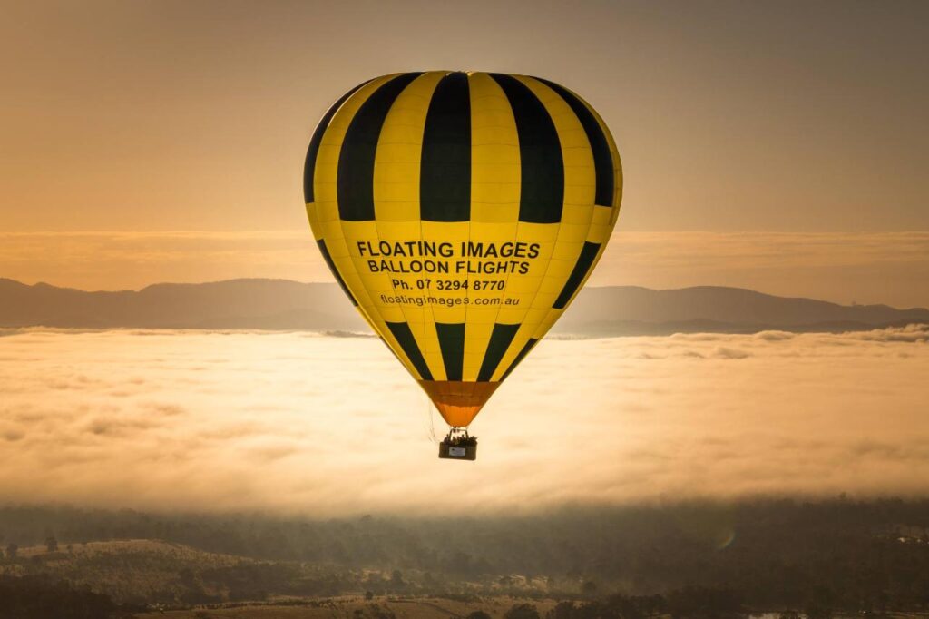 A yellow and black hot air balloon with "Floating Images Balloon Flights" printed on it flies above clouds at sunrise, with hills visible in the background.