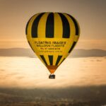 A yellow and black hot air balloon with "Floating Images Balloon Flights" printed on it flies above clouds at sunrise, with hills visible in the background.