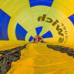 View from inside a yellow and blue hot air balloon envelope, looking toward the opening where equipment and people are visible preparing for inflation.