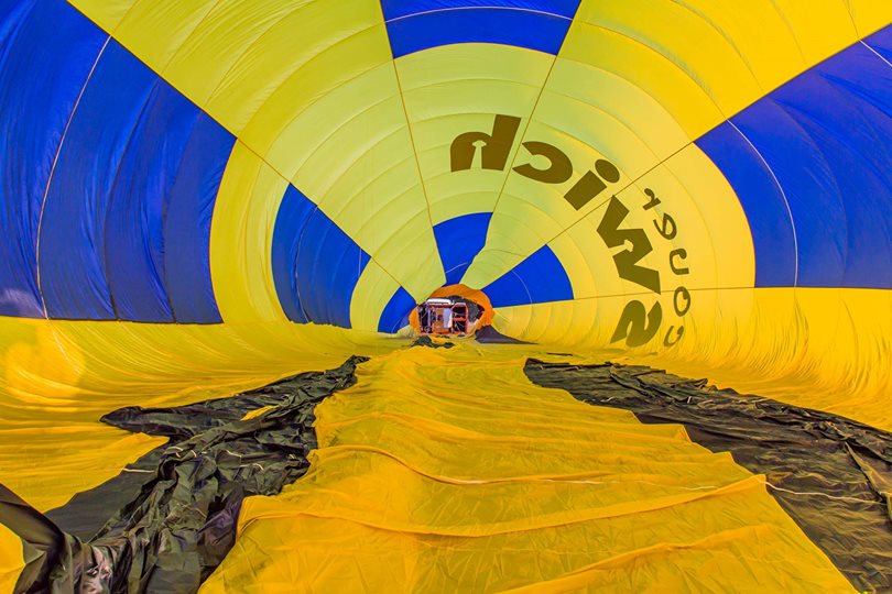 View from inside a yellow and blue hot air balloon envelope, looking toward the opening where equipment and people are visible preparing for inflation.
