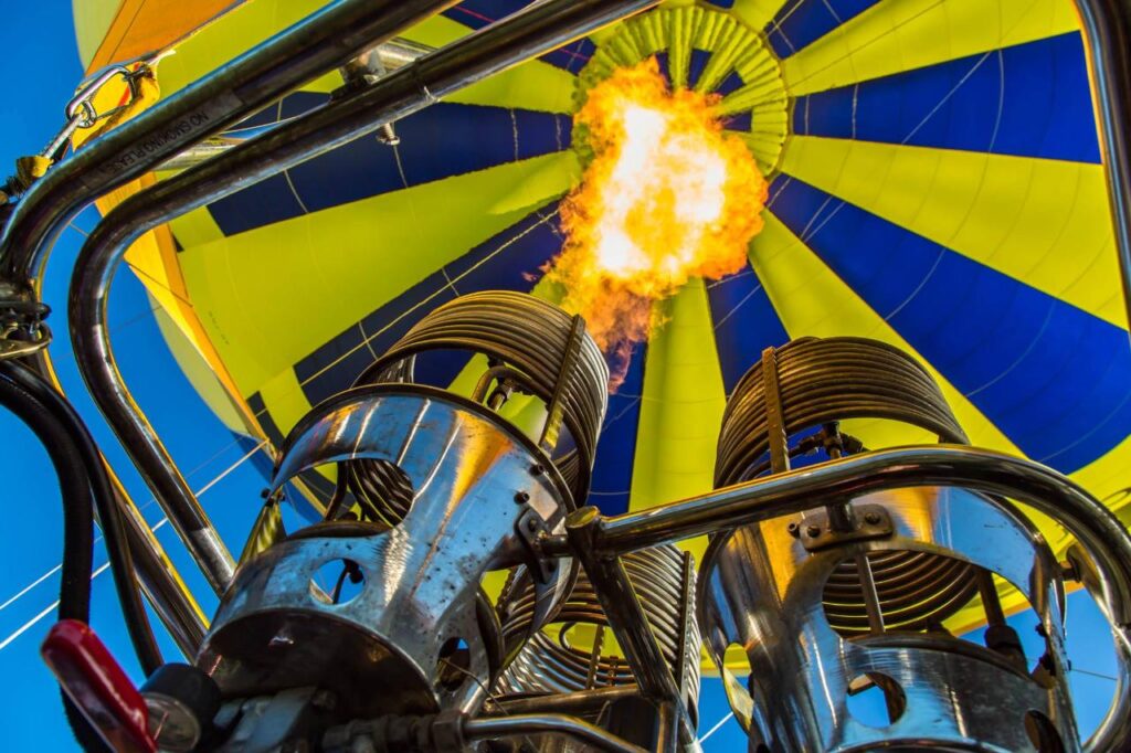 Close-up view from below of a hot air balloon burner releasing flames upward into the yellow and blue balloon canopy.