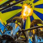 Close-up view from below of a hot air balloon burner releasing flames upward into the yellow and blue balloon canopy.