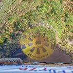 View from a hot air balloon basket looking down at the ground, with the balloon’s shadow visible on a grassy and partially wet landscape.