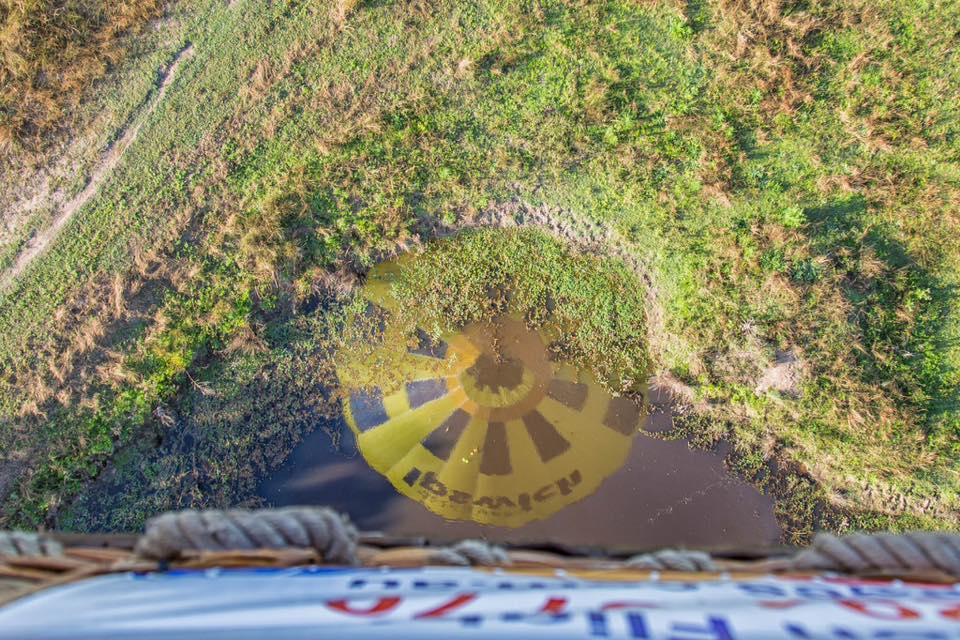 View from a hot air balloon basket looking down at the ground, with the balloon’s shadow visible on a grassy and partially wet landscape.