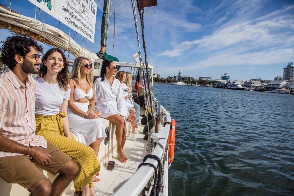 A group of people sit and relax on a sailboat in sunny weather, with a city waterfront and blue sky visible in the background.