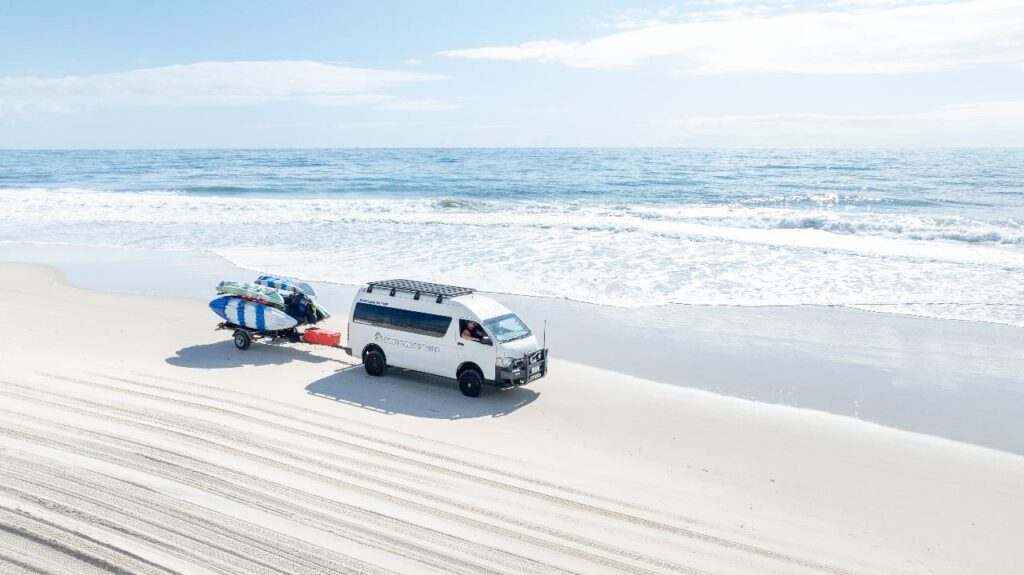 A white van with a trailer carrying several surfboards is parked on a sandy beach near the ocean under a partly cloudy sky.