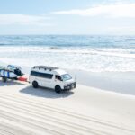 A white van with a trailer carrying several surfboards is parked on a sandy beach near the ocean under a partly cloudy sky.
