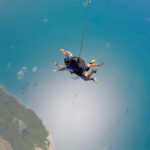 A person skydiving over a coastline, suspended by a parachute harness above blue ocean water and land below.