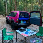 A pink SUV with its back open is parked at a wooded campsite. Camping chairs, a table with cooking supplies, and storage bins are set up nearby on the leaf-covered ground.