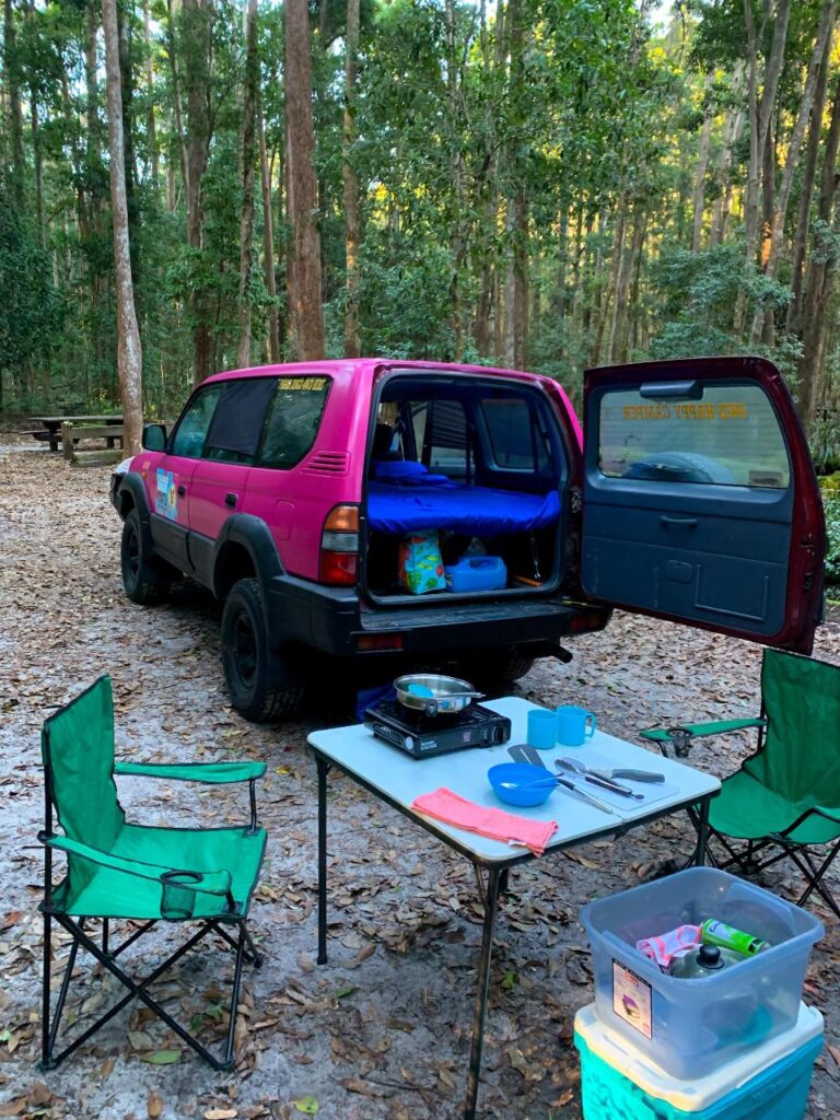 A pink SUV with its back open is parked at a wooded campsite. Camping chairs, a table with cooking supplies, and storage bins are set up nearby on the leaf-covered ground.